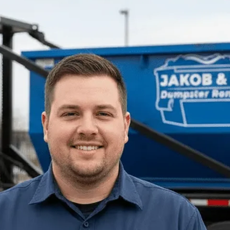 Ryan Voss - Lead Driver Ryan Voss, Lead Driver at Jakob & Sons Dumpster Rental Flint, standing in front of a company rolloff dumpster